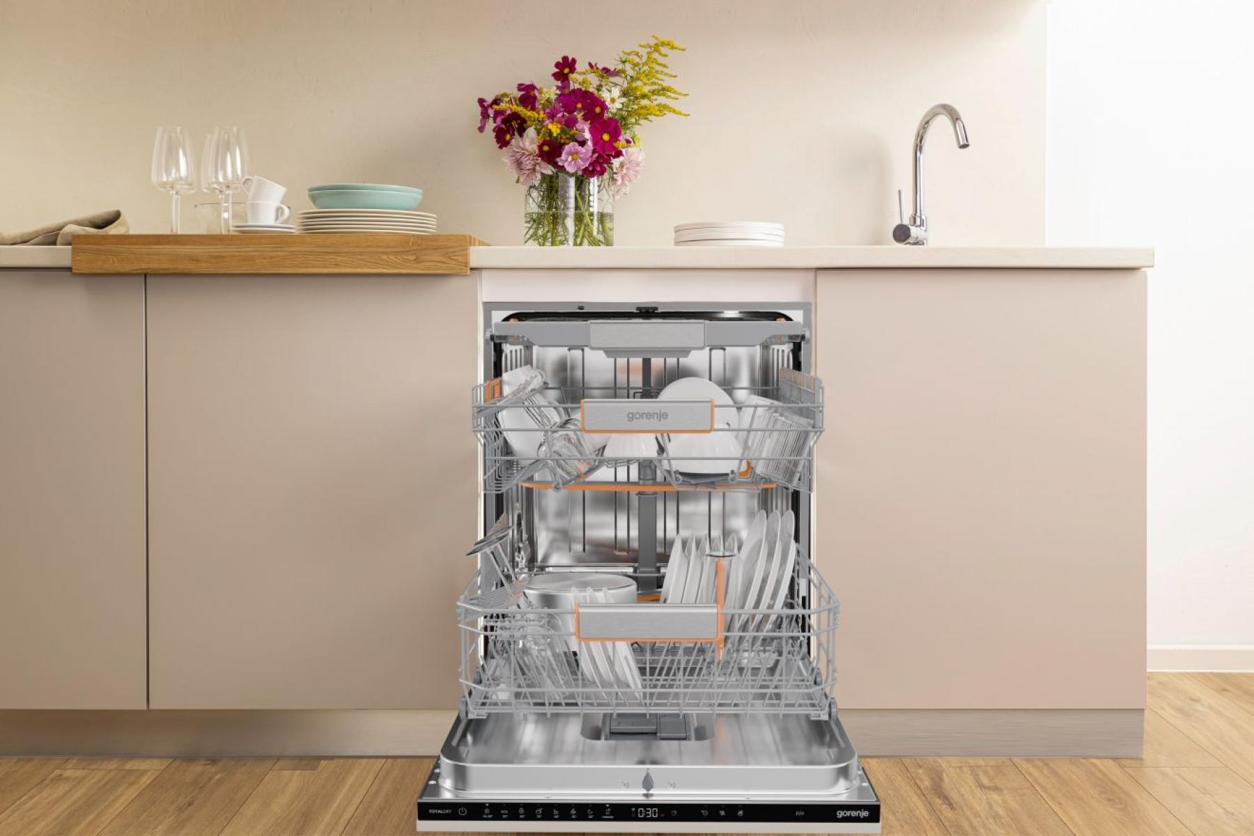 Otwarta zmywarka wypełniona naczyniami. Kwiaty, szklanki i talerze na blacie. Beżowe szafki i drewniana podłoga.