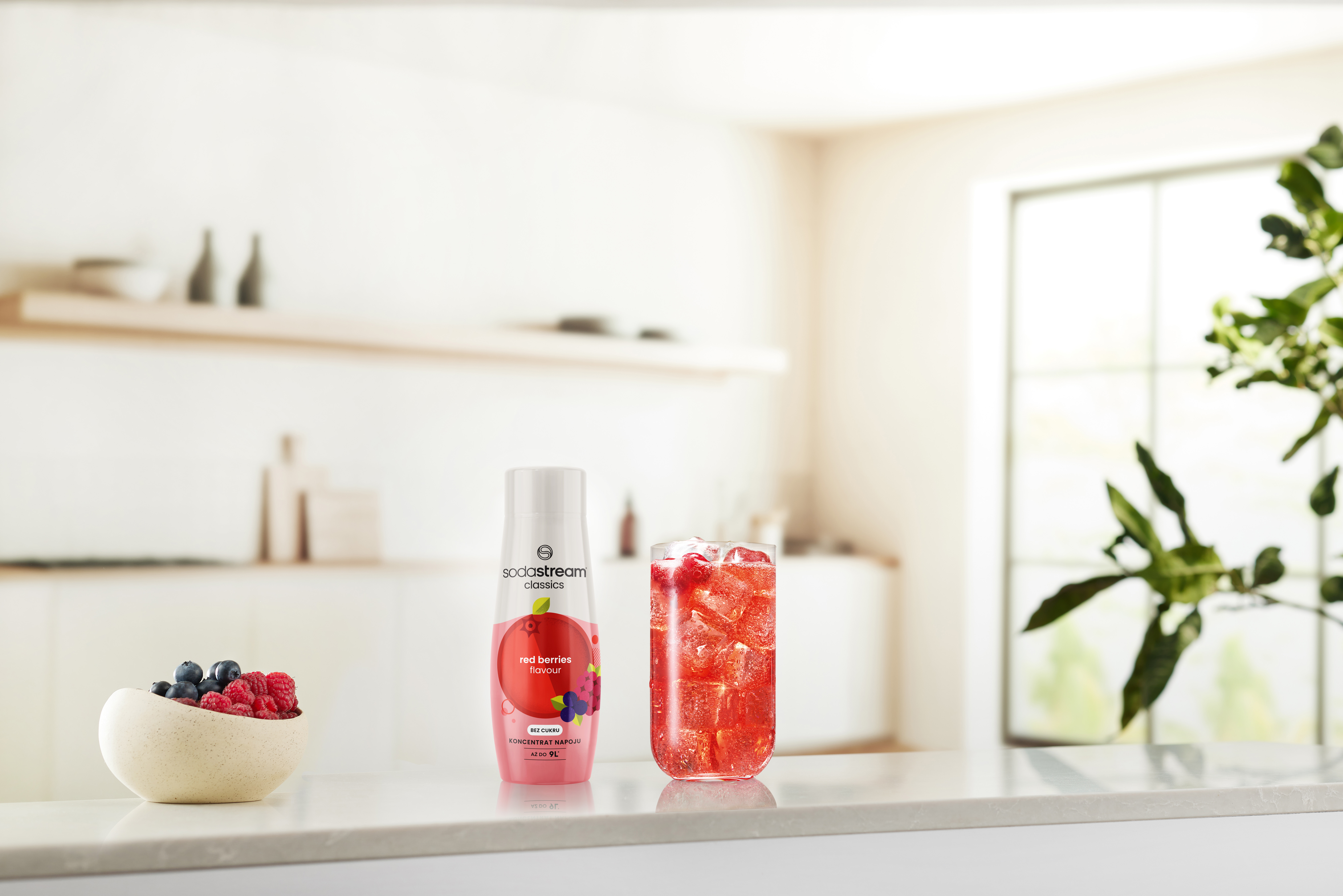 Na blacie szklanka z napojem z czerwonych owoców, butelka i miska z jagodami. W tle kuchnia.