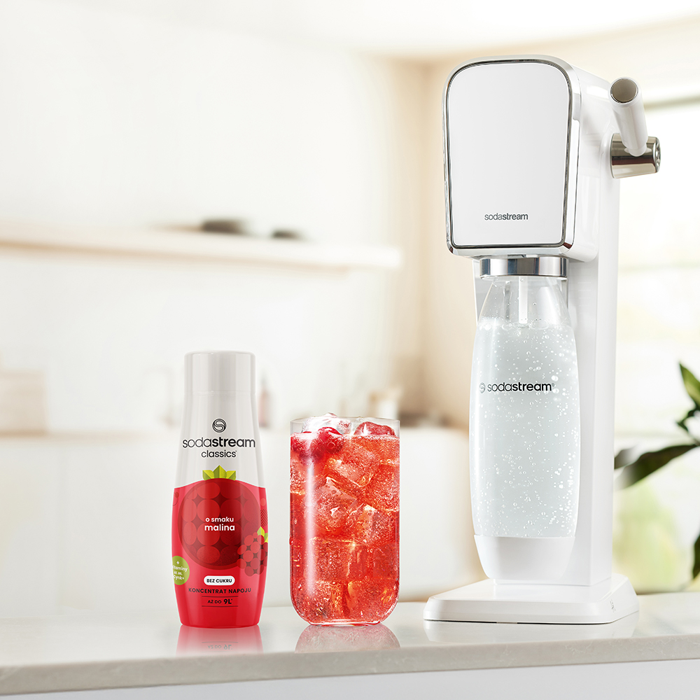 Białe urządzenie SodaStream, butelka syropu malinowego i szklanka napoju malinowego na białym blacie, rozmyte tło kuchni.