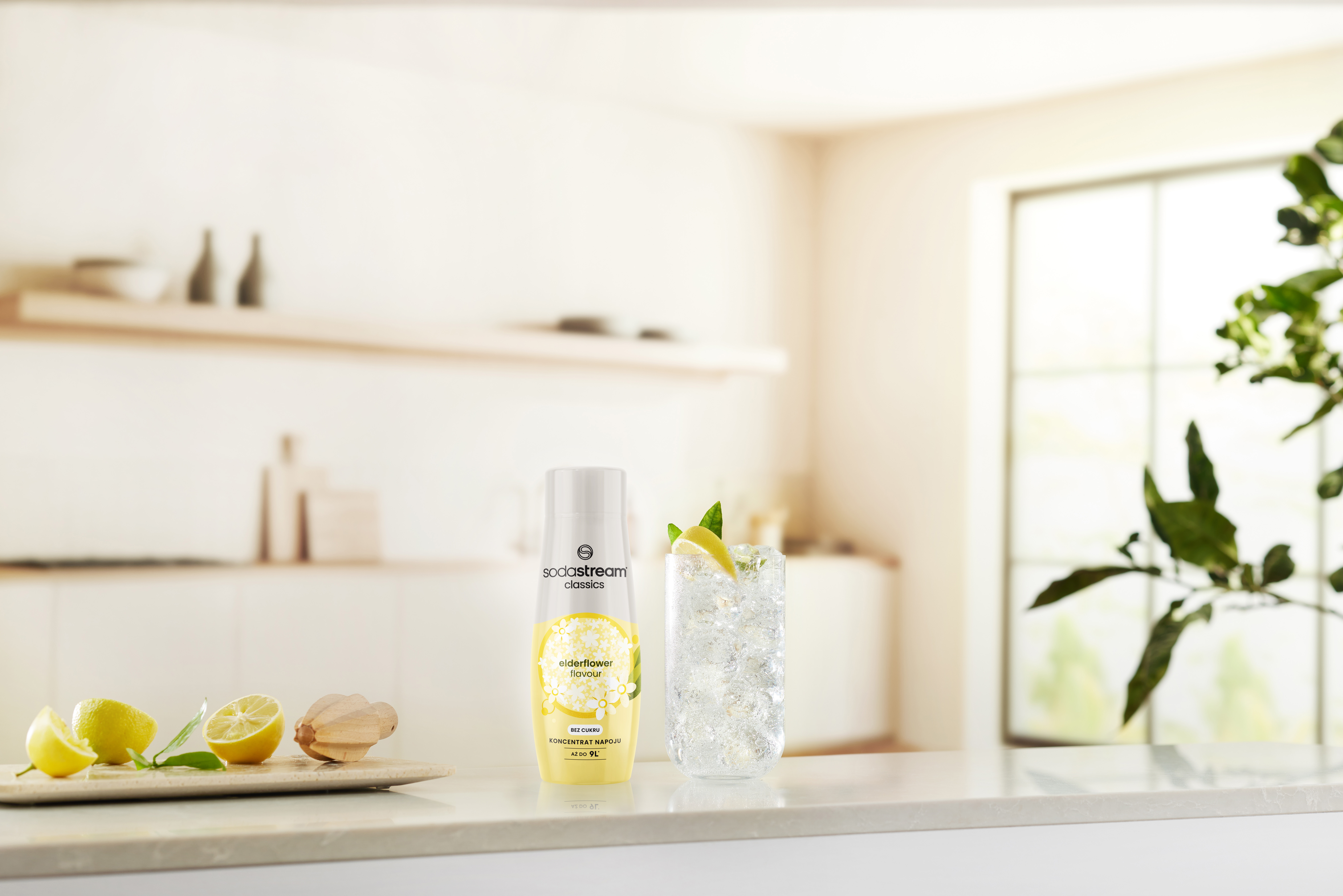 Butelka SodaStream, szklanka lemoniady z lodem i cytryną oraz cytryny na desce do krojenia stoją na blacie.