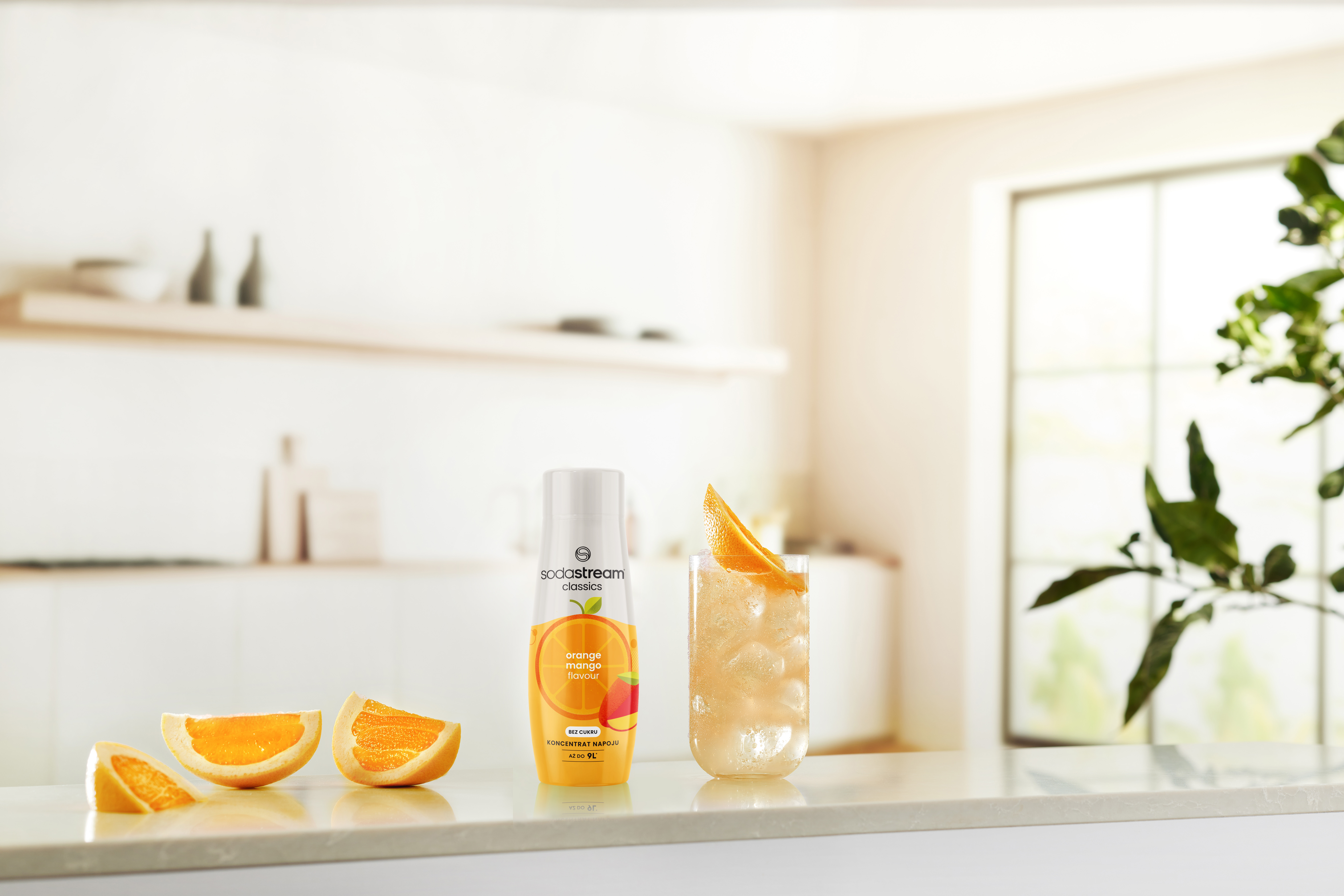 Butelka SodaStream, napój, plasterki pomarańczy i szklanka sody z lodem leżą na białym blacie. Tło jest rozmyte.