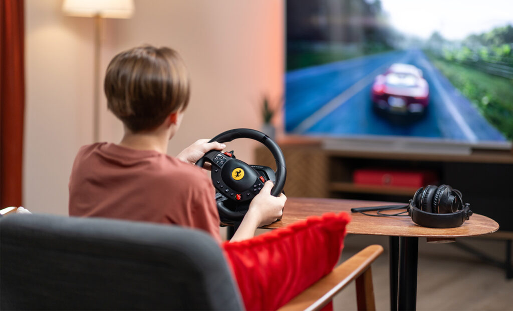 Une personne joue à un jeu de course avec un volant. Une Ferrari rouge à l'écran. Casque sur la table.