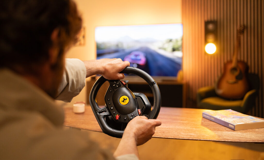 Une personne tenant un volant Ferrari, jouant à un jeu de course. Une guitare, un fauteuil et un livre sont en arrière-plan.