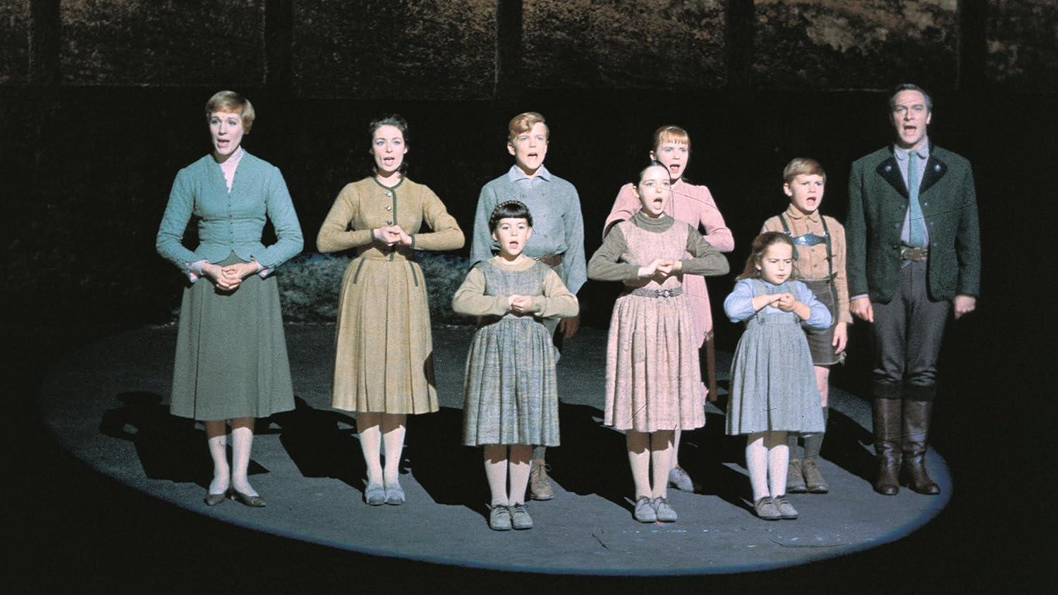 Eine Gruppe von Menschen, darunter Erwachsene und Kinder, steht auf einer Bühne. Sie singen mit verschiedenen Ausdrücken vor einem dunklen Hintergrund.
