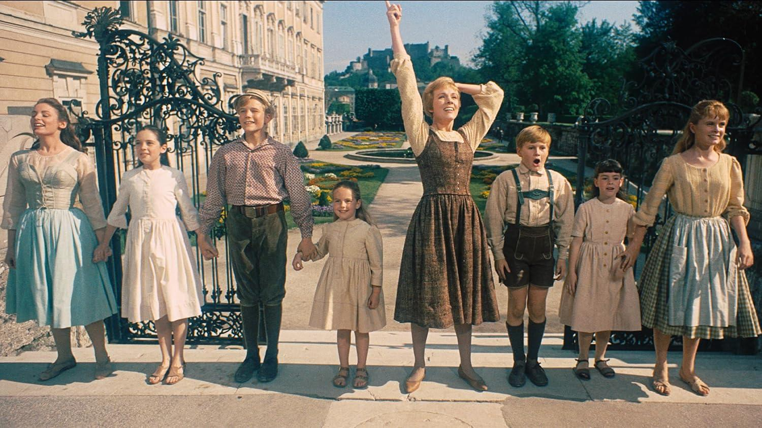 Eine Gruppe von Menschen, darunter Kinder, hält Hände und lächelt. Eine Frau in der Mitte hebt den Arm. Im Hintergrund ist ein Gebäude zu sehen.