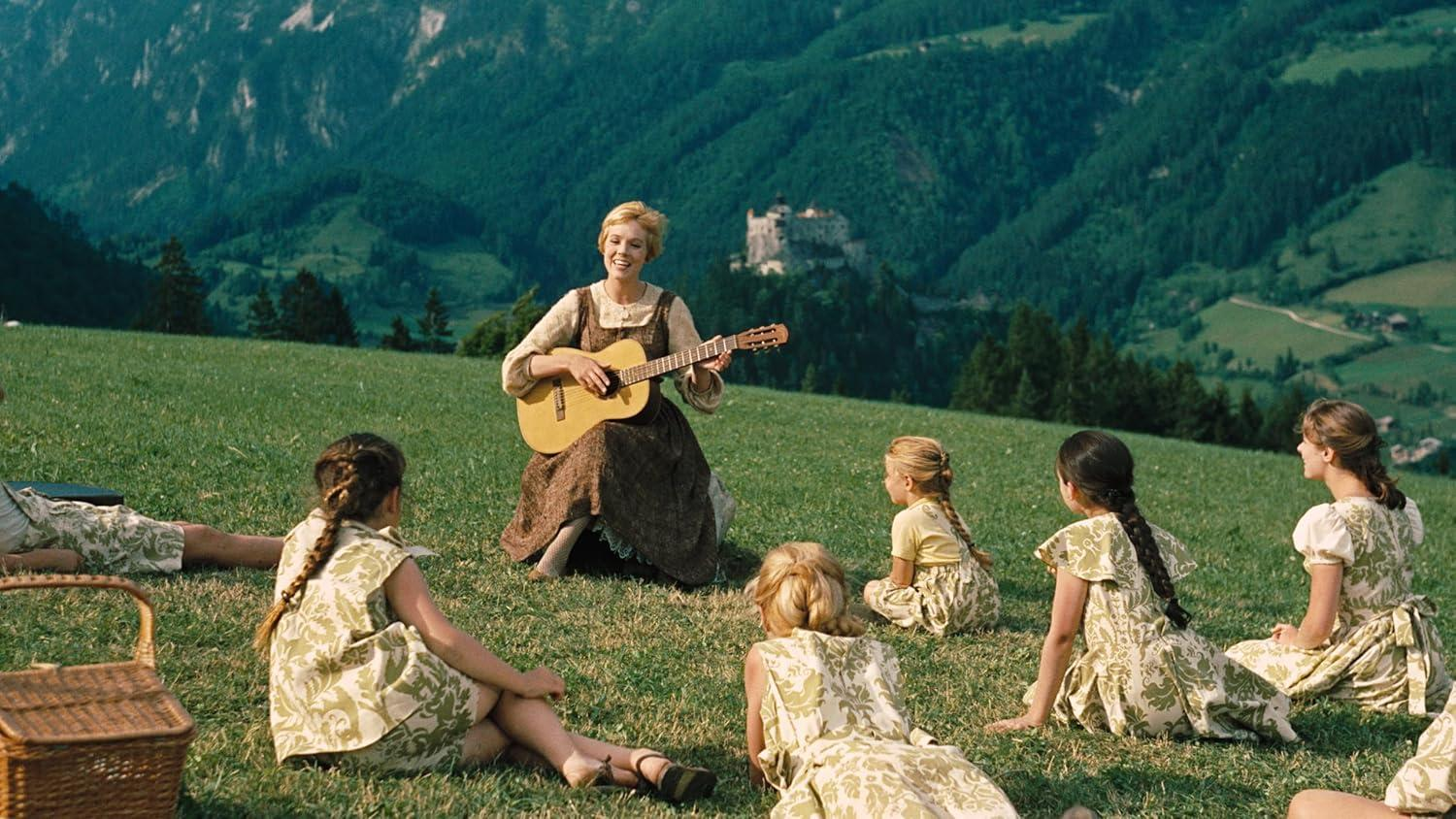 Eine Frau spielt Gitarre, umgeben von Kindern auf einem grasbewachsenen Hügel. Berge und ein Schloss sind im Hintergrund.