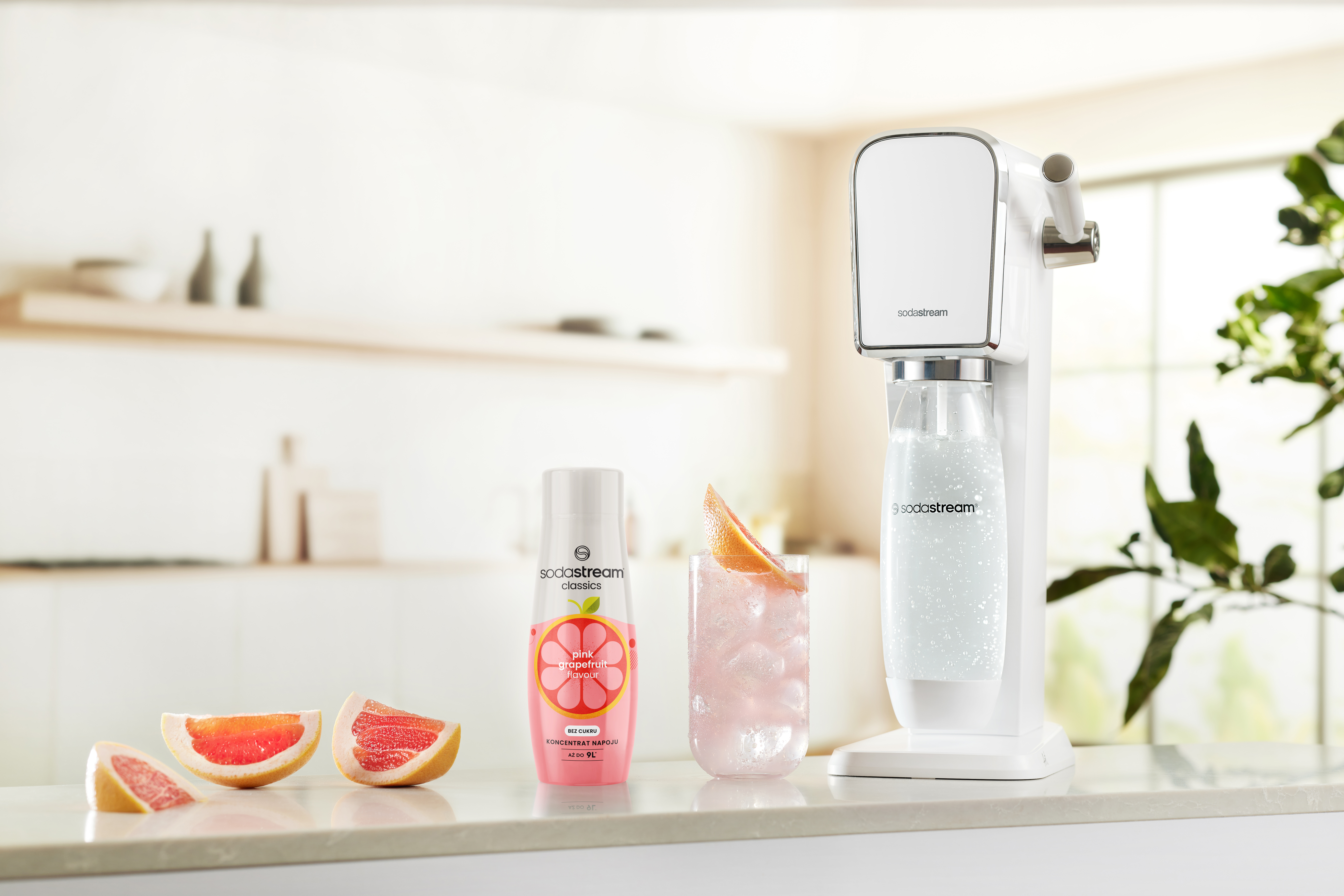 Automat SodaStream, plasterki grejpfruta, różowy napój z lodem i butelka różowego syropu grejpfrutowego na blacie.