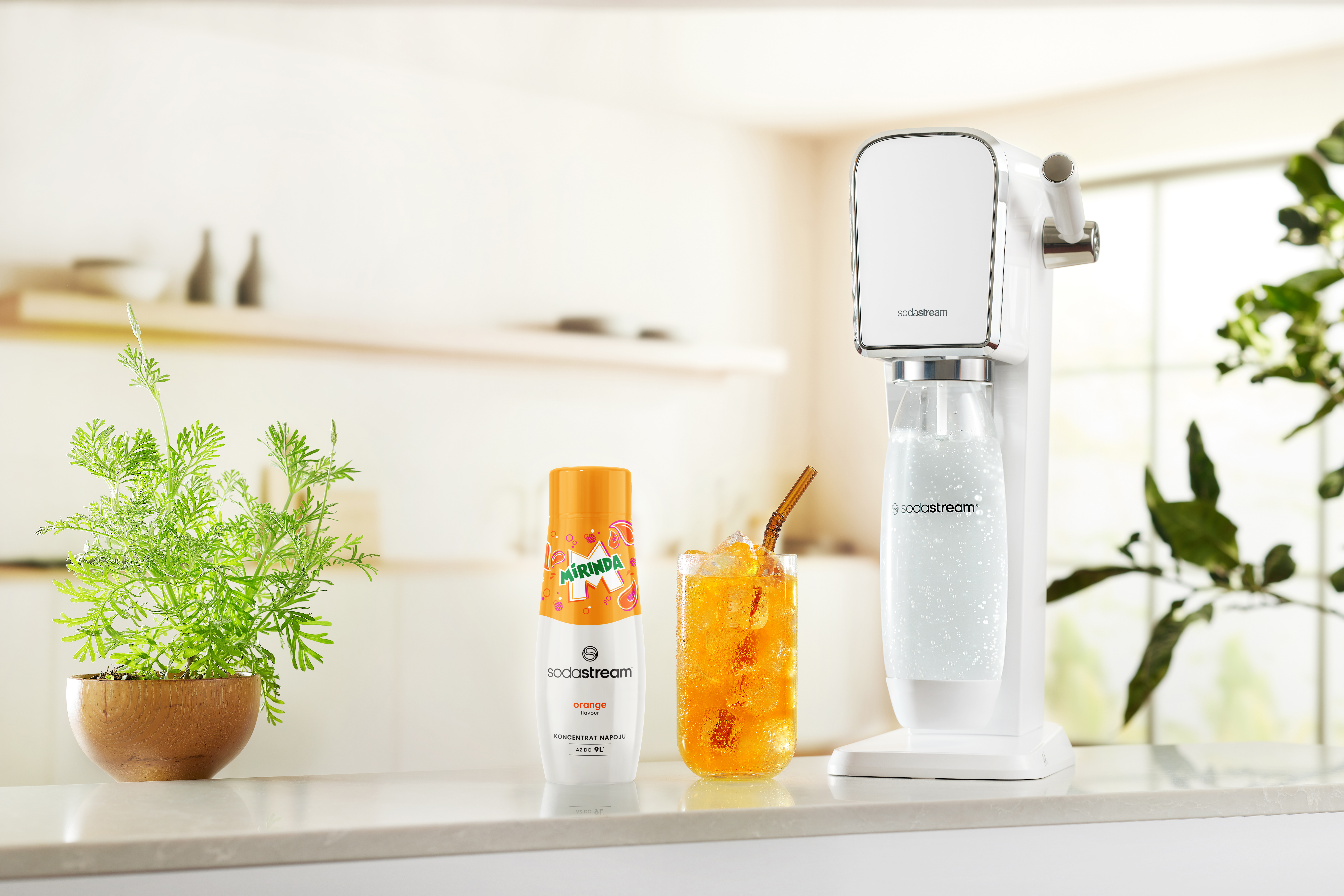 Na blacie stoi automat SodaStream, pomarańczowa soda i butelka Mirindy. Roślina jest na pierwszym planie.