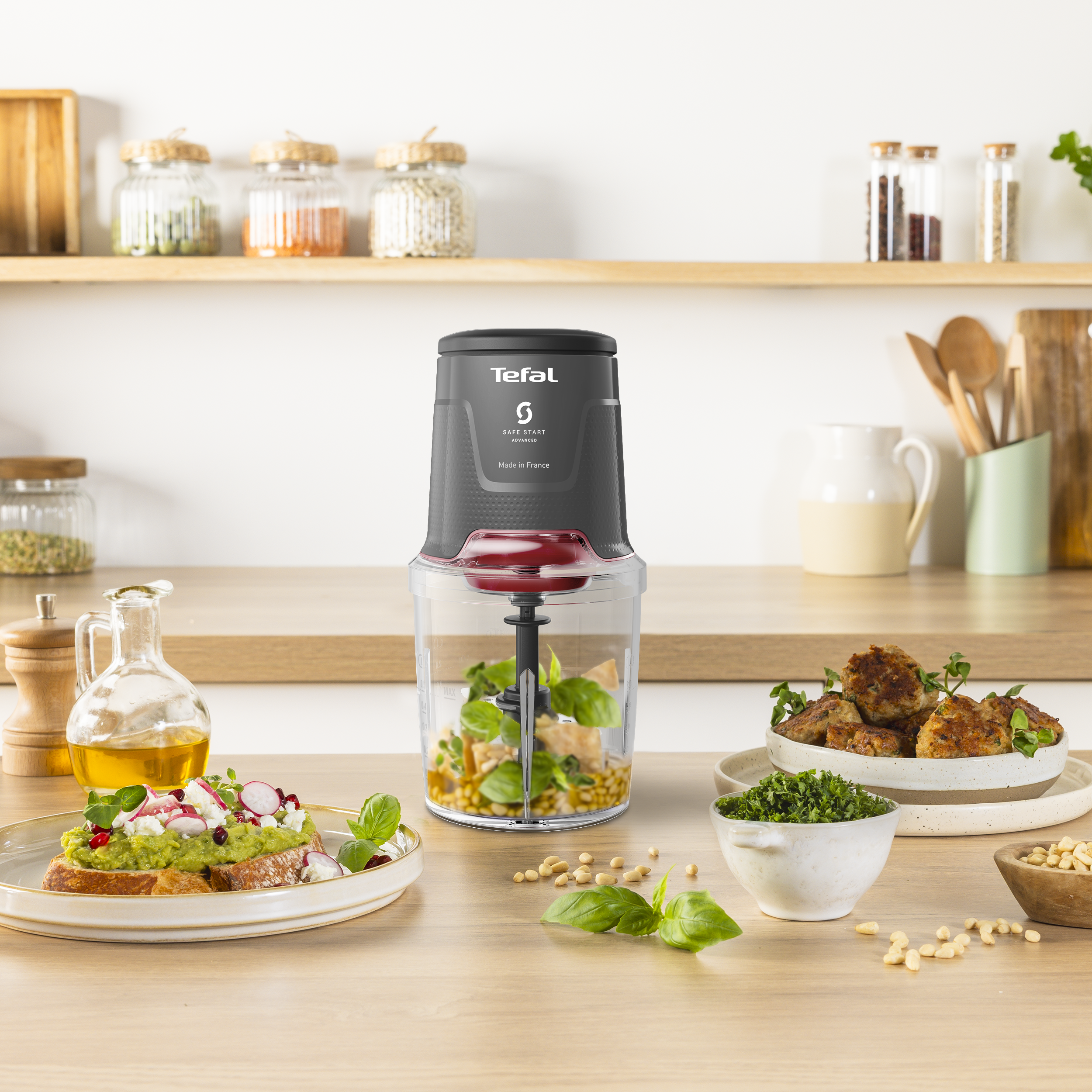 Egy élelmiszer-feldolgozó egy faasztalon van, olyan élelmiszerekkel, mint a pesto és az avokádós kenyér. A háttérben üvegek vannak.