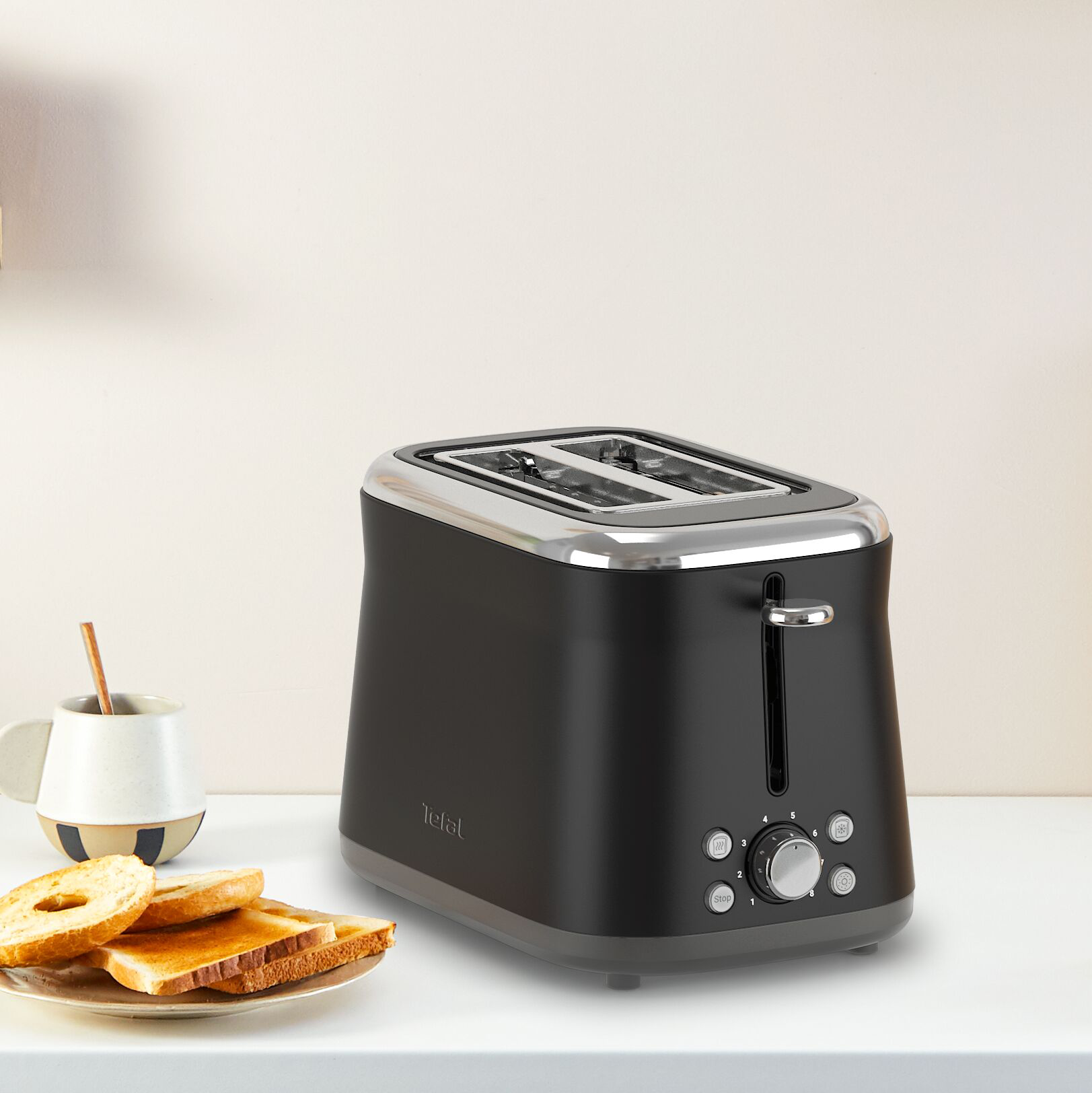 Egy fekete Tefal kenyérpirító bagel-lel és pirítóssal. Egy csésze kanállal balra. A háttér világos bézs.