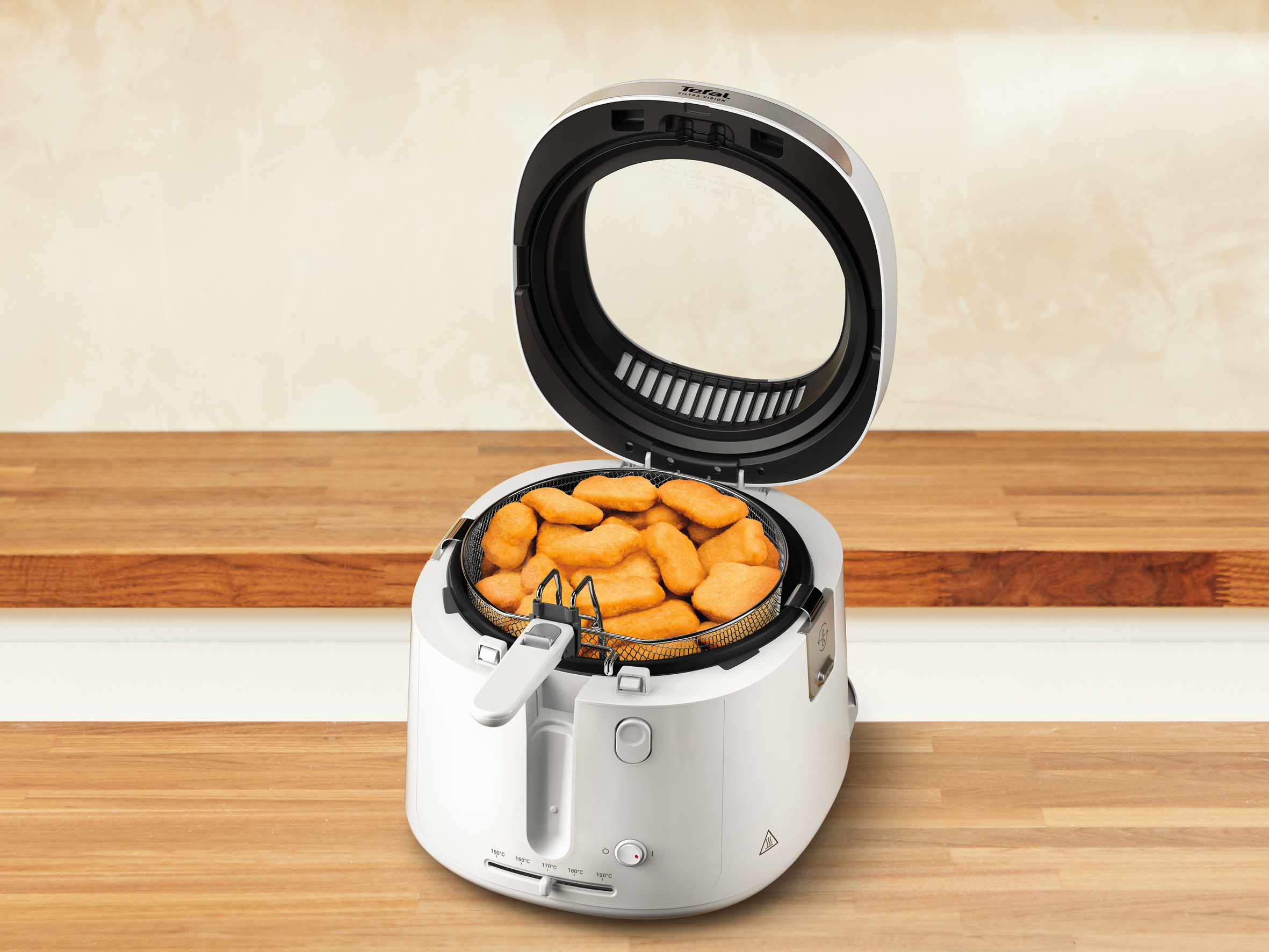 Egy fehér Tefal légkeveréses sütő, benne nuggetsekkel egy kosárban. Fa felületen van, világos bézs háttérrel.
