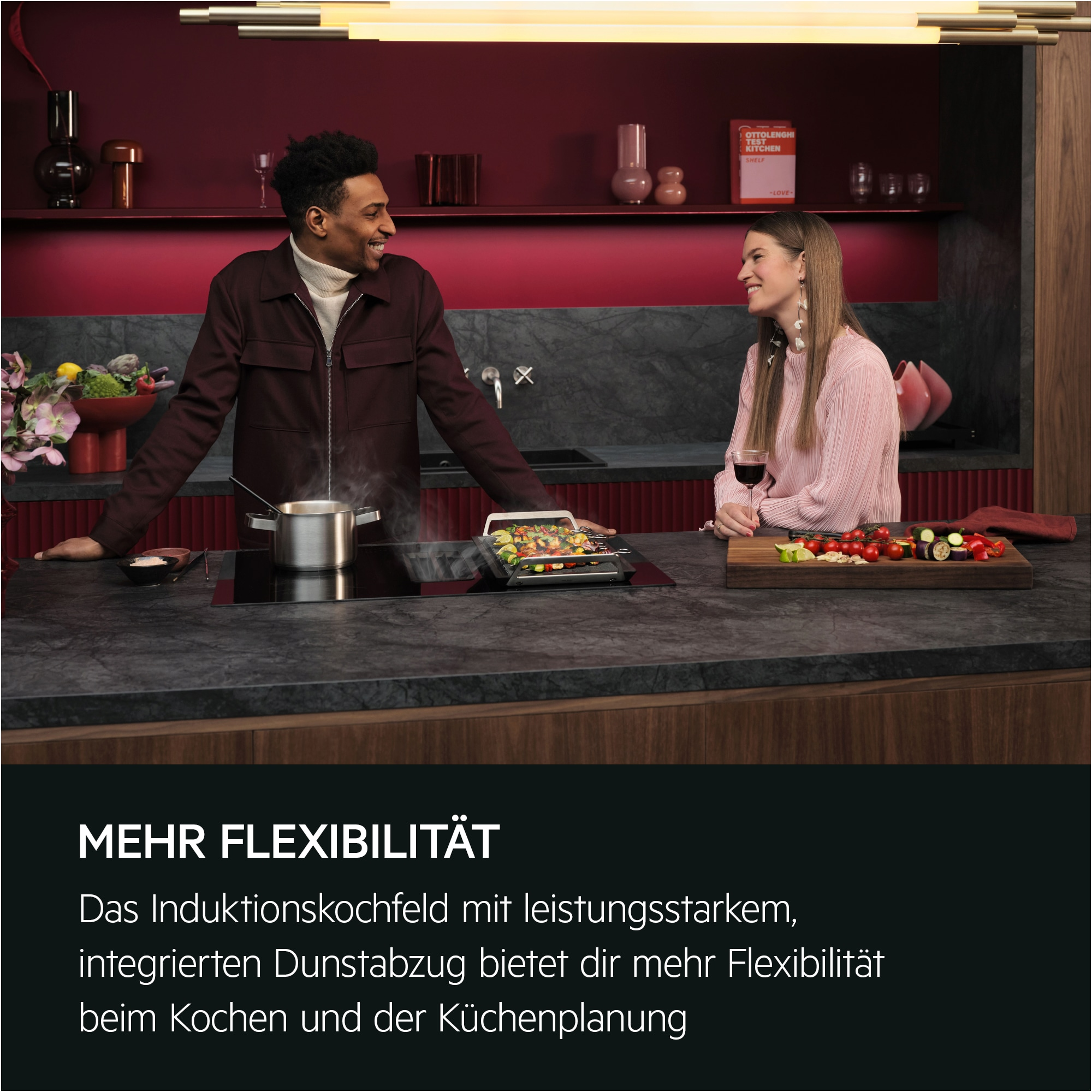 Ein Paar lächelt in einer Küche. Ein Topf dampft auf einem Kochfeld. Zutaten und ein Kochbuch liegen auf der Theke.