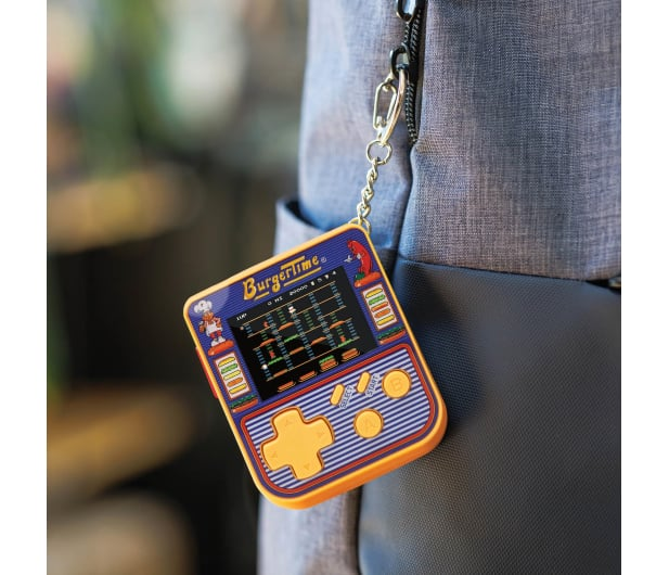 Przenośna gra BurgerTime wisi na breloczku. Jest pomarańczowa i niebieska, z małym ekranem i przyciskami sterowania, przymocowana do szarej torby.