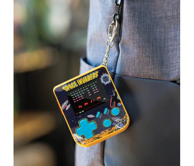 Gra przenośna Space Invaders wisi na breloczku. Gra jest żółta i niebieska, przymocowana do szarego plecaka.