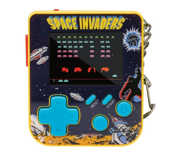 Przenośna gra Space Invaders. Ma żółtą ramkę, niebieski pad kierunkowy i ekran wyświetlający grę. Jest przymocowana do metalowego łańcucha.