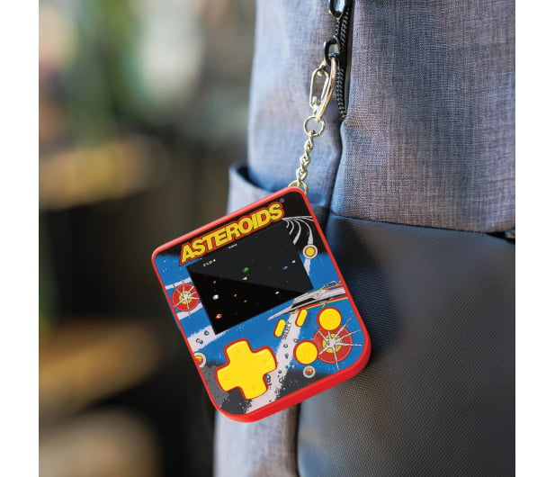 Przenośna konsola do gier z grą Asteroids jest przymocowana do breloka. Konsola wisi na szarym plecaku osoby.