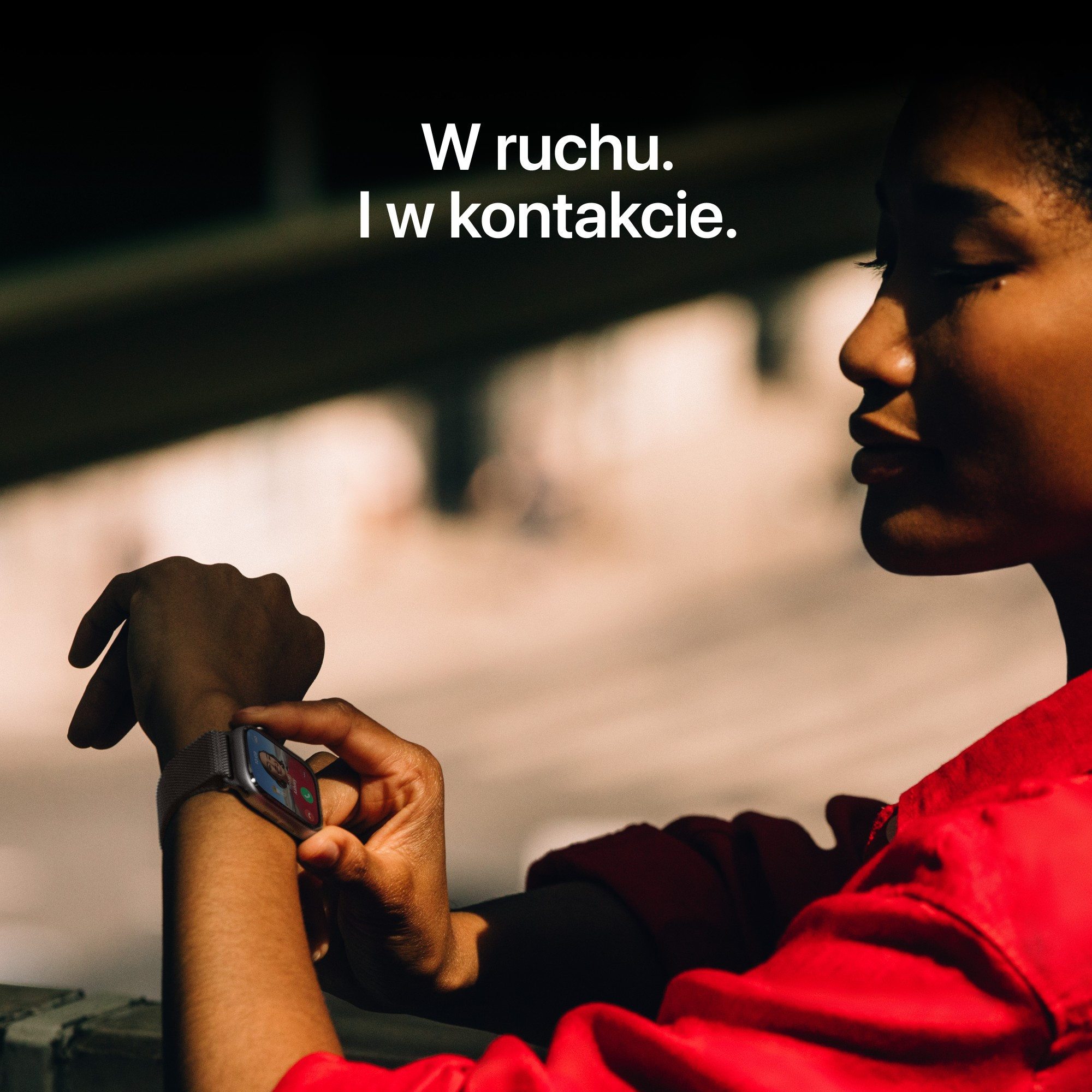Kobieta w czerwonej kurtce spogląda na swój smartwatch. Tekst na zdjęciu: 'W ruchu, I w kontakcie'.