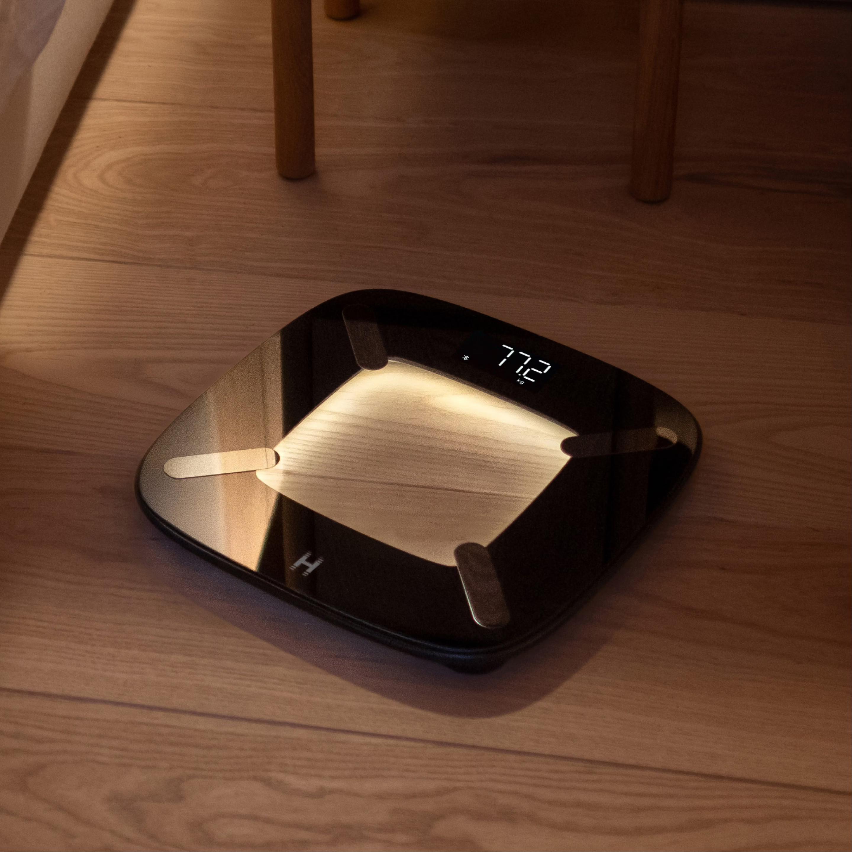 Czarna inteligentna waga pokazuje 77,2 kg na drewnianej podłodze. Waga jest blisko łóżka i krzesła.