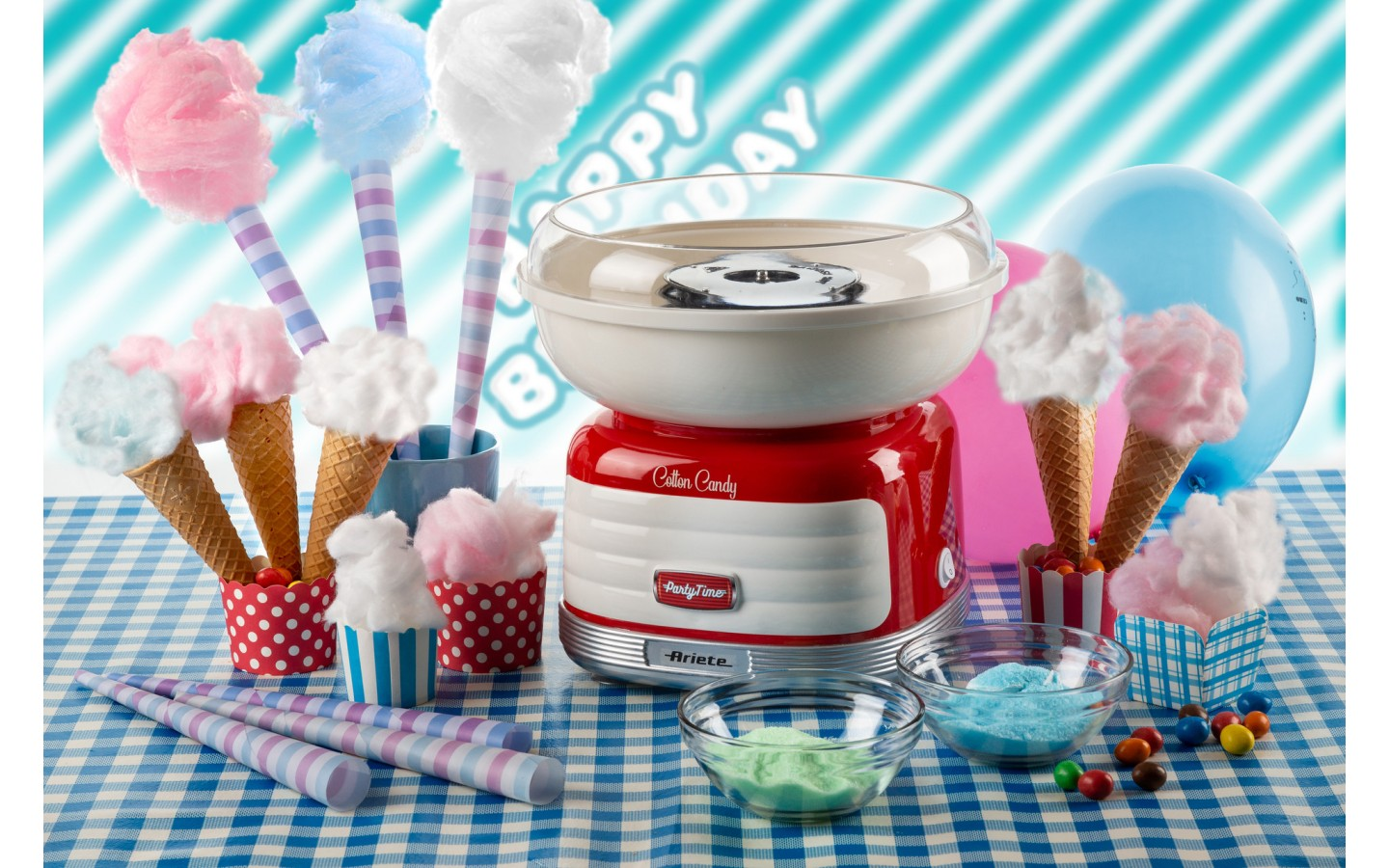 Maszyna do waty cukrowej z różnymi kolorowymi smakołykami, rożkami i cukierkami na kraciastej serwetce. Balony w tle.
