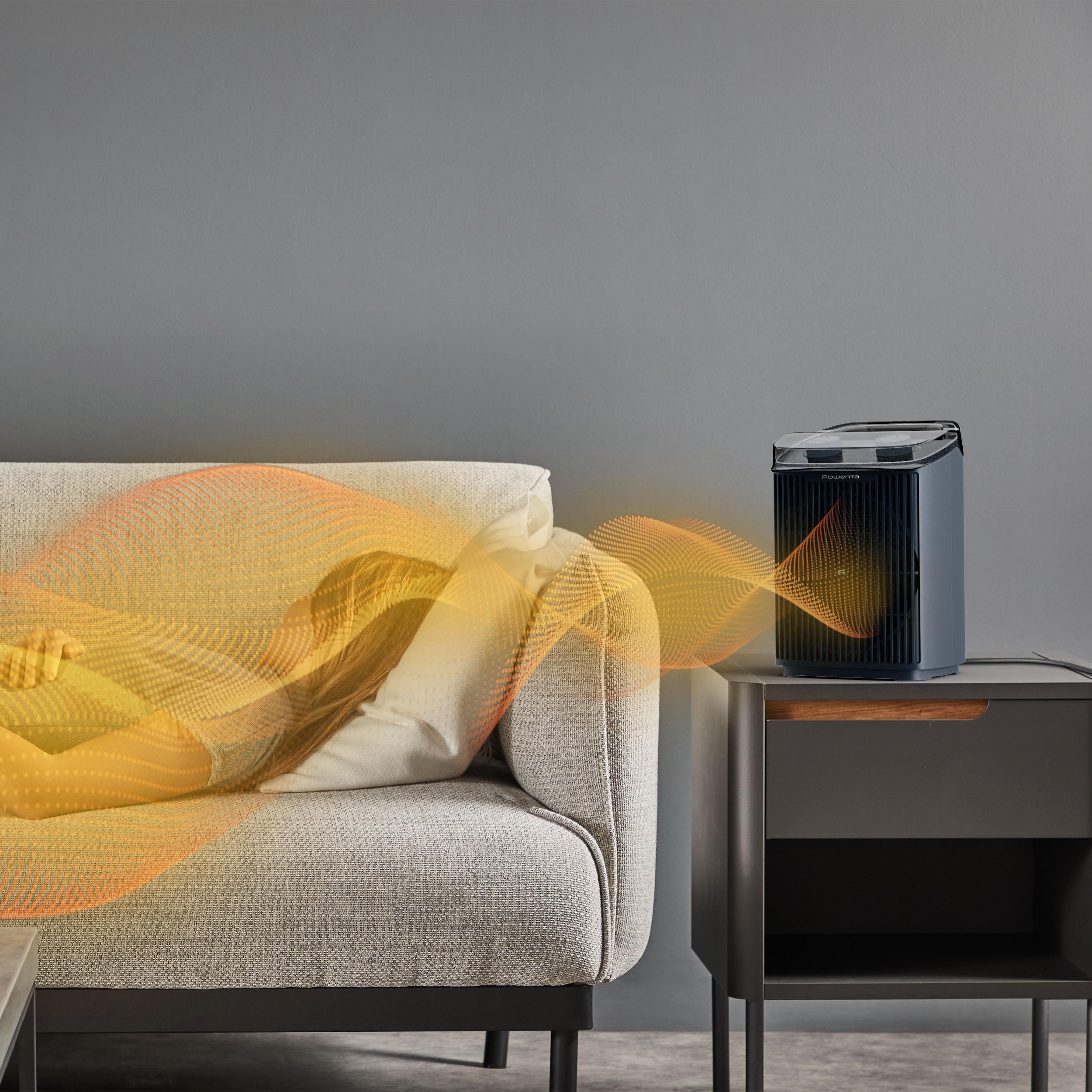 Eine Person liegt auf einem Sofa neben einem dunkelgrauen Heizgerät. Orangefarbene Wellen gehen vom Heizgerät auf die Person aus.