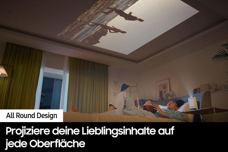 Ein Projektor zeigt Bilder an der Decke. Zwei Personen liegen auf einem Bett und schauen sich die Bilder an. Ein Raum mit Fenstern und Vorhängen.