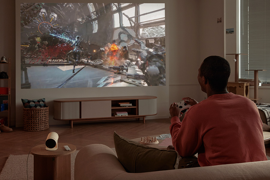 Ein Mann spielt ein Videospiel, das auf eine Wand projiziert wird. Das Spiel zeigt eine Kampfszene. Er hält einen Controller.