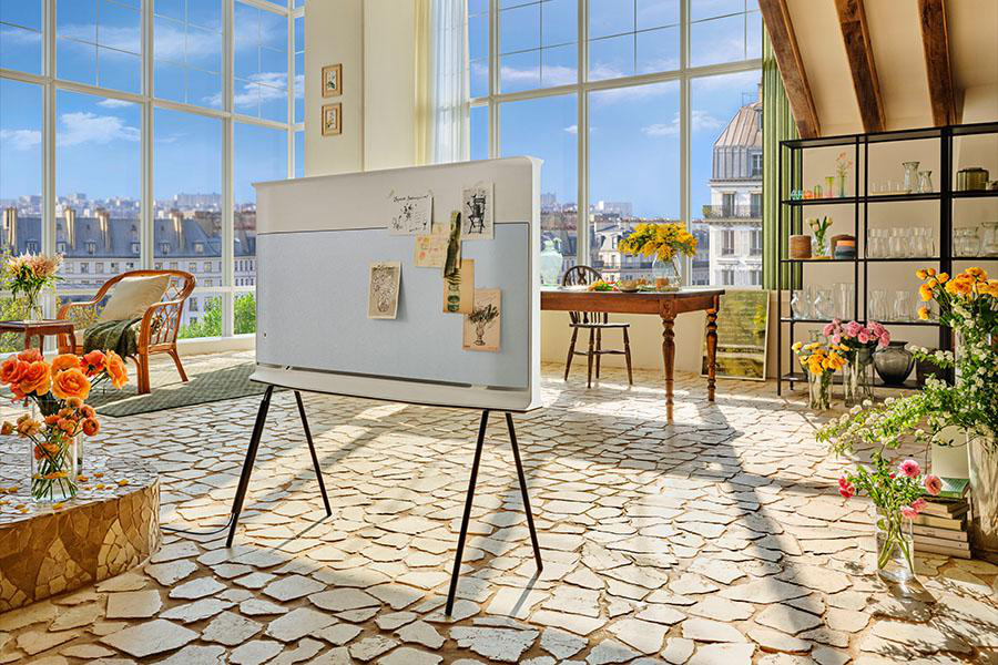Wohnzimmer mit Fernseher auf Ständer, große Fenster mit Stadtblick. Blumen in Vasen. Steinplattenboden. Tisch und Stühle.