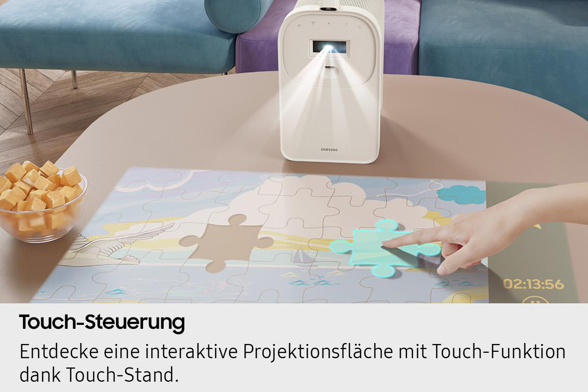 Ein weißer Projektor zeigt ein Puzzle an. Eine Hand berührt ein Puzzleteil. Würfel in einer Schüssel auf dem Tisch.