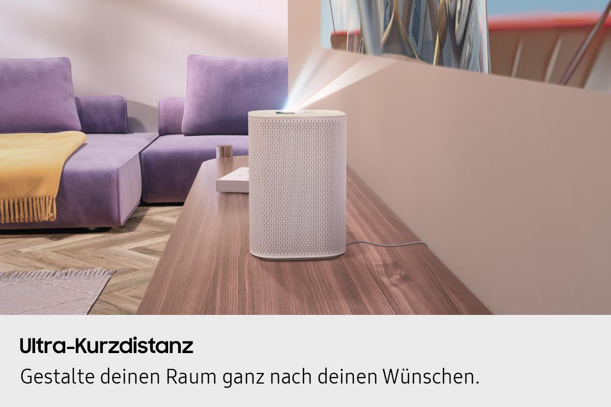 Ein weißer Lautsprecher auf einem Holztisch projiziert Licht. Lila Couch und gelbe Decke im Hintergrund.