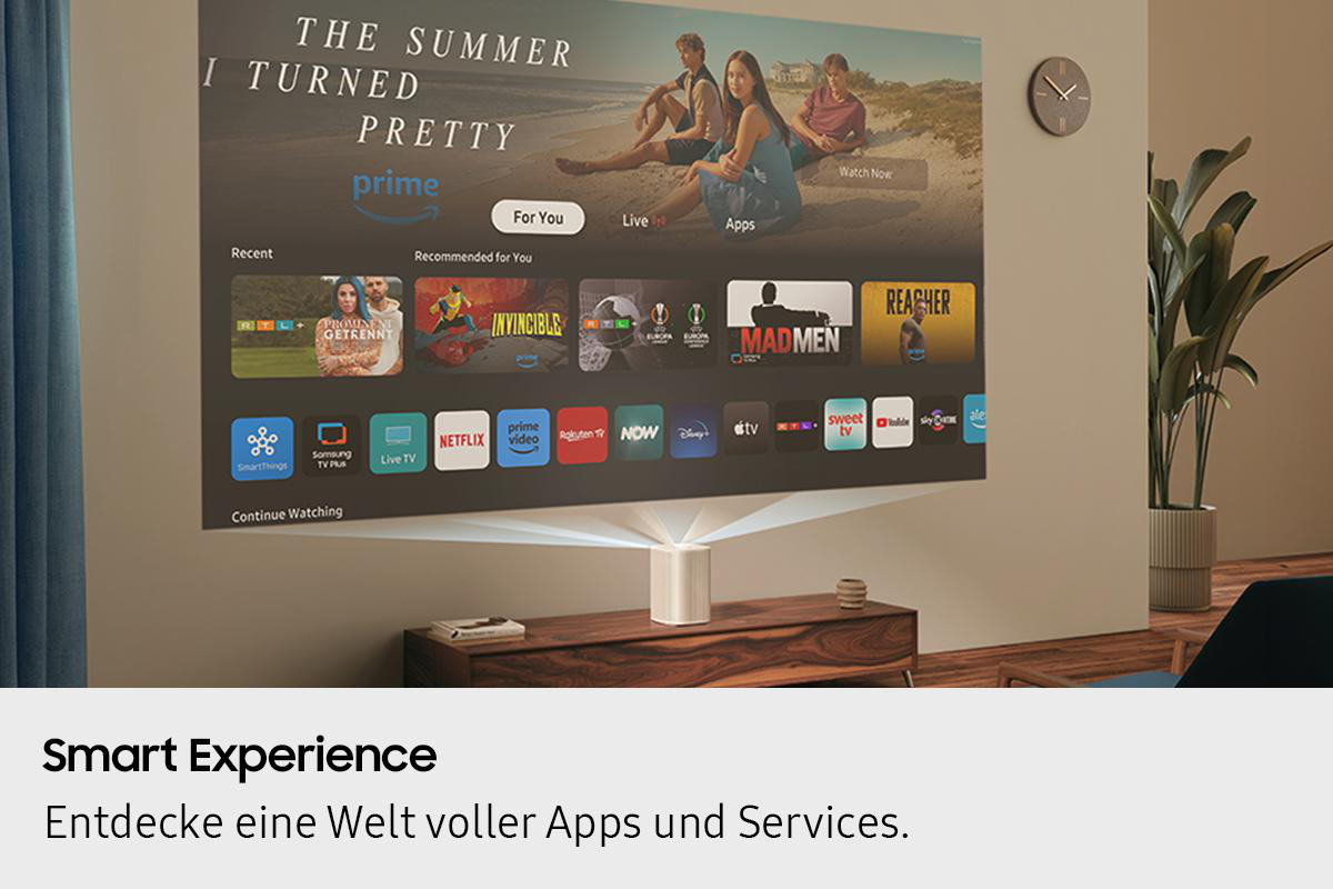 Ein Projektor zeigt eine TV-Oberfläche. Eine Uhr hängt an der Wand. Im Hintergrund ist eine Pflanze, davor ein Holzschrank.