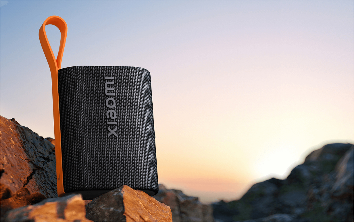 Czarny głośnik z logo Xiaomi, pomarańczowym paskiem, na skałach z tłem zachodu słońca.