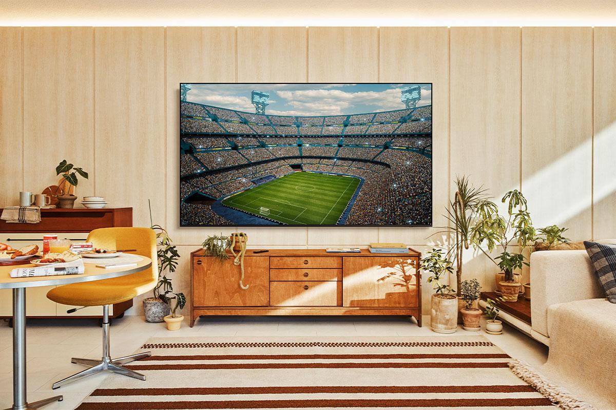 Ein Wohnzimmer mit einem großen Fernseher, der ein Fußballstadion zeigt. Es gibt einen Tisch, Pflanzen, einen Teppich und einen Holzschrank.
