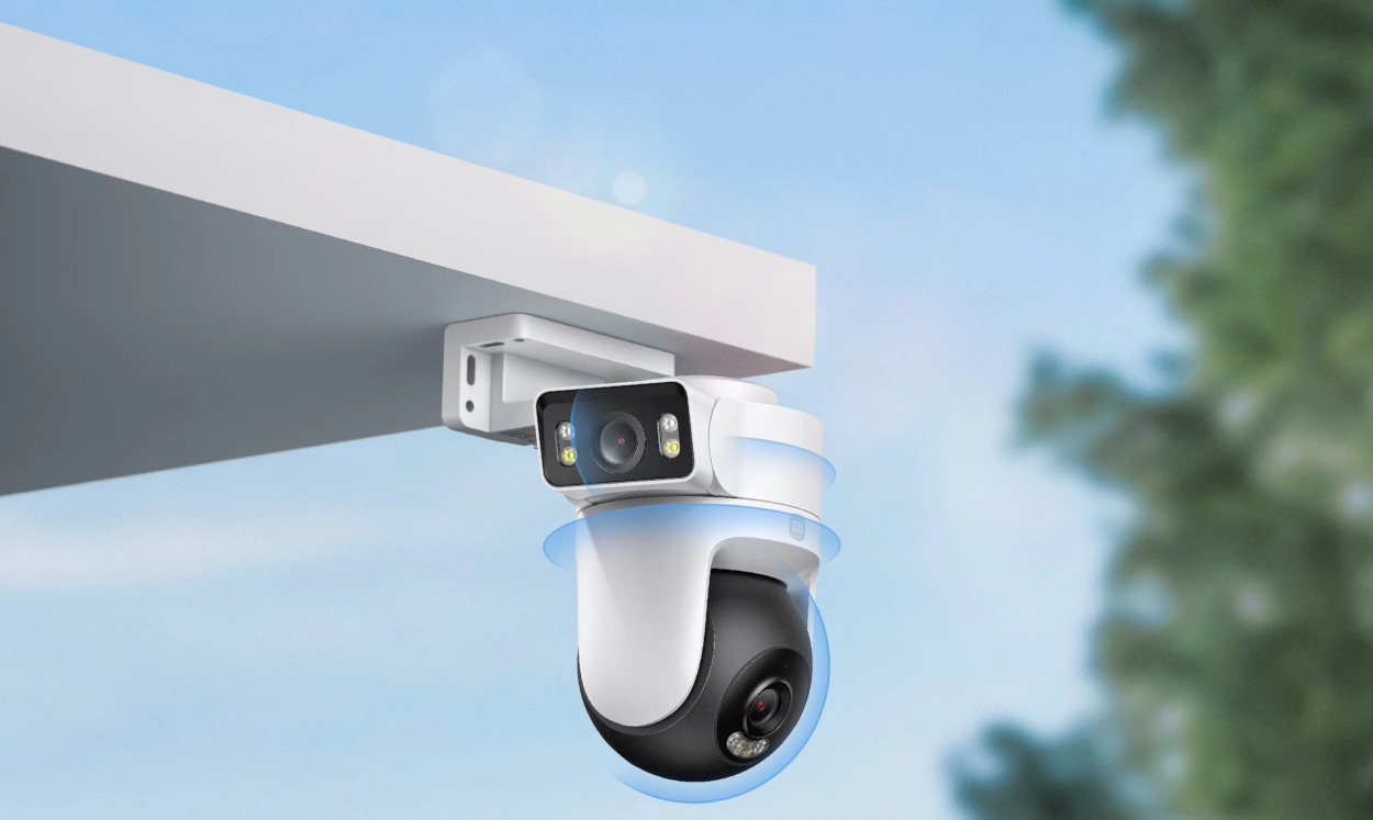 Biała i czarna kamera bezpieczeństwa jest zamontowana na białej powierzchni, na tle błękitnego nieba z zielonymi liśćmi.