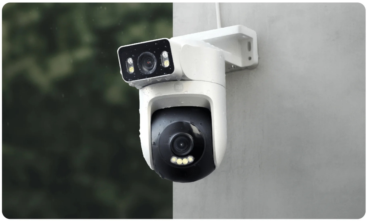 Biała kamera bezpieczeństwa jest zamontowana na ścianie. Ma dwa obiektywy i jest otoczona rozmytym zielonym tłem.