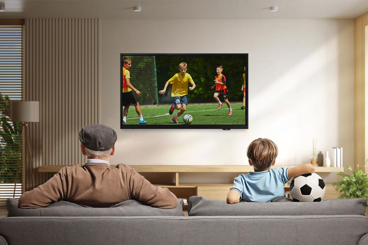 Ein Mann und ein Junge sehen sich ein Fußballspiel im Fernsehen an. Ein Fußball liegt auf der Couch. Das TV zeigt Kinder beim Fußballspielen.
