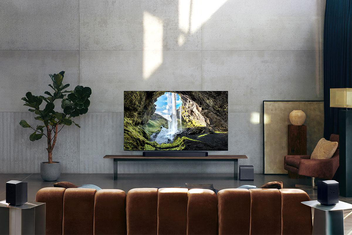 Ein modernes Wohnzimmer mit großem Fernseher, der eine Wasserfallszene zeigt. Braunes Sofa, Lautsprecher und eine Pflanze im Raum.