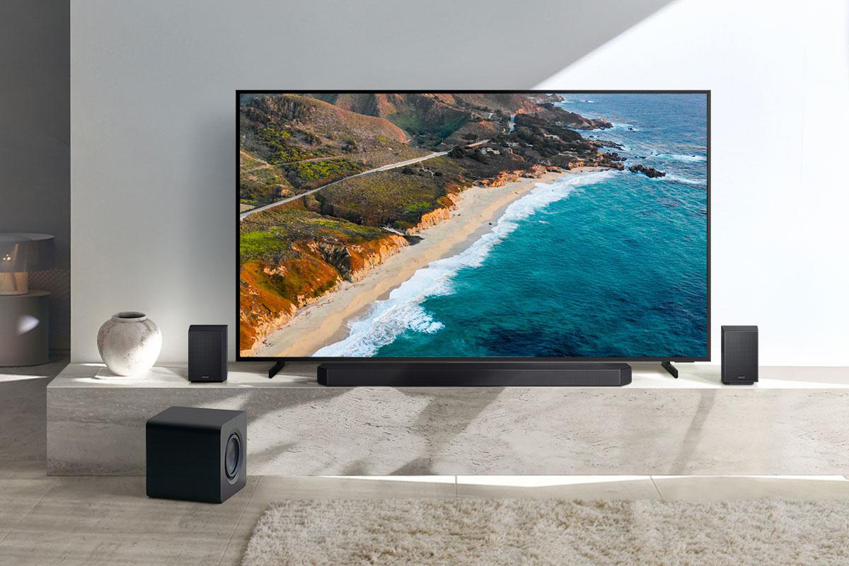 Ein großer Fernseher zeigt eine Strandszene. Sound-System-Komponenten befinden sich darunter und an den Seiten. Ein Subwoofer steht davor.