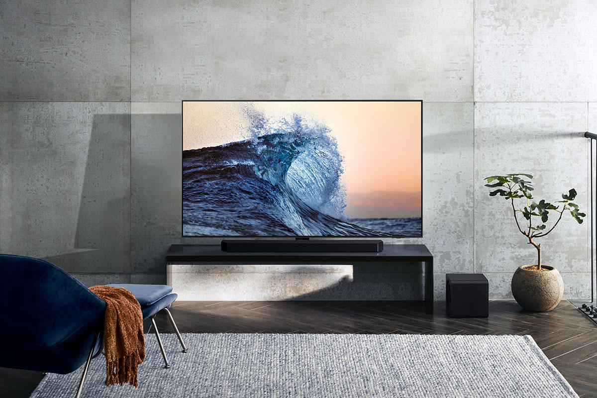 Ein großer Fernseher zeigt eine Welle. Darunter befindet sich eine Soundbar. Davor steht ein blauer Stuhl mit einer Decke. Rechts ist eine Pflanze.