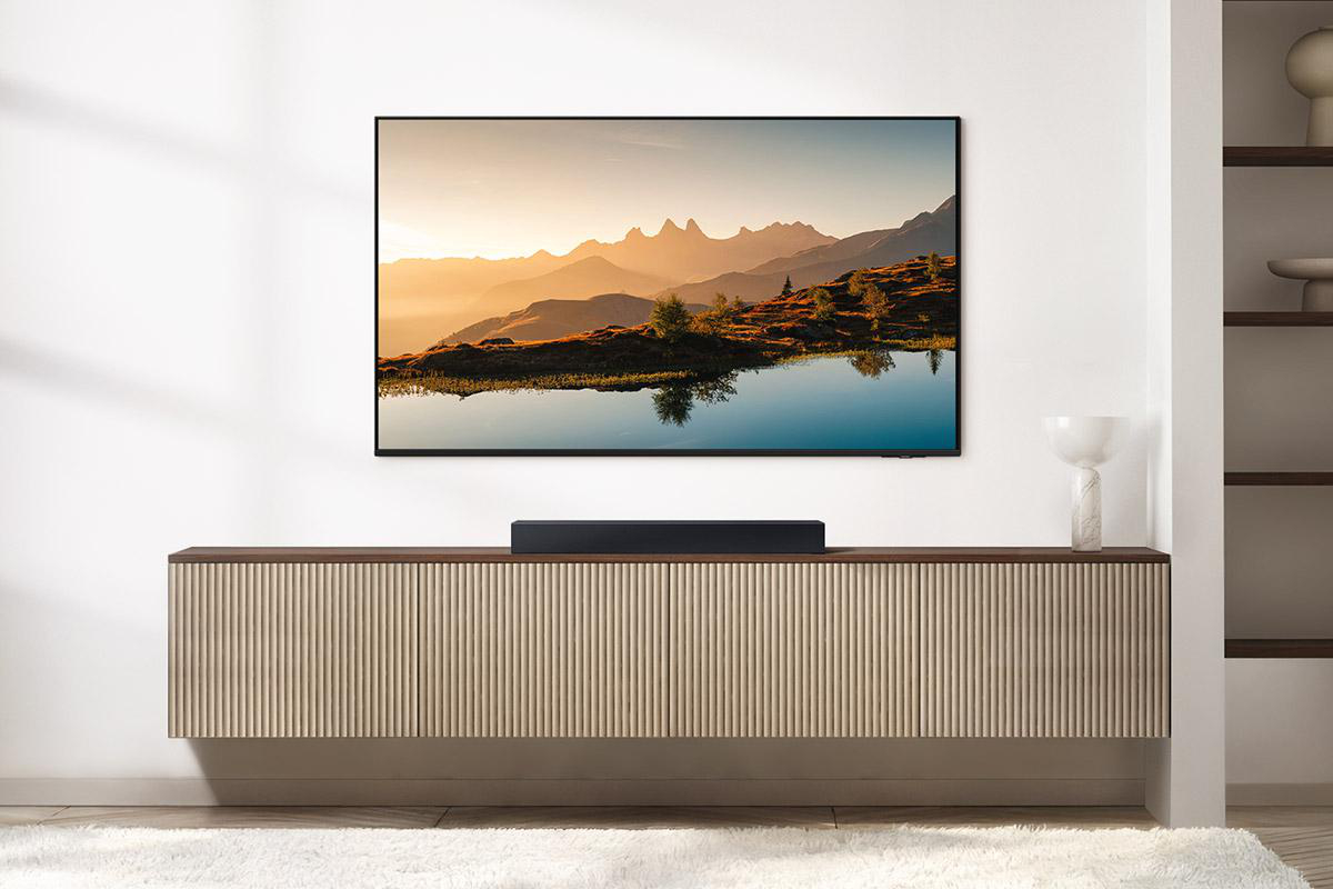 Ein Fernseher zeigt eine Bergseeszene. Darunter befindet sich eine Soundbar auf einem Holzschrank. Der Raum hat eine weiße Wand und einen Teppich.