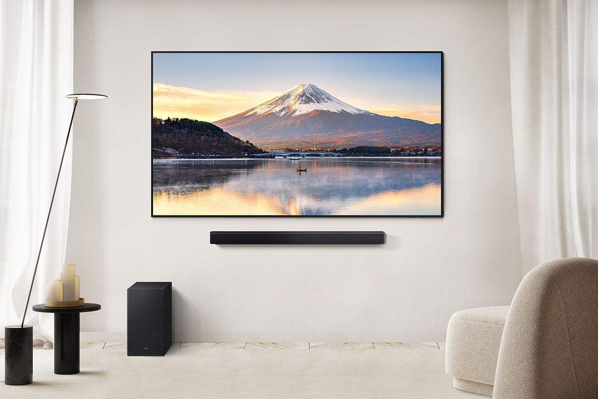 Ein Fernseher zeigt eine Berglandschaft über Wasser. Darunter eine Soundbar. Ein Subwoofer, eine Lampe, ein Tisch und ein Stuhl befinden sich im Raum.