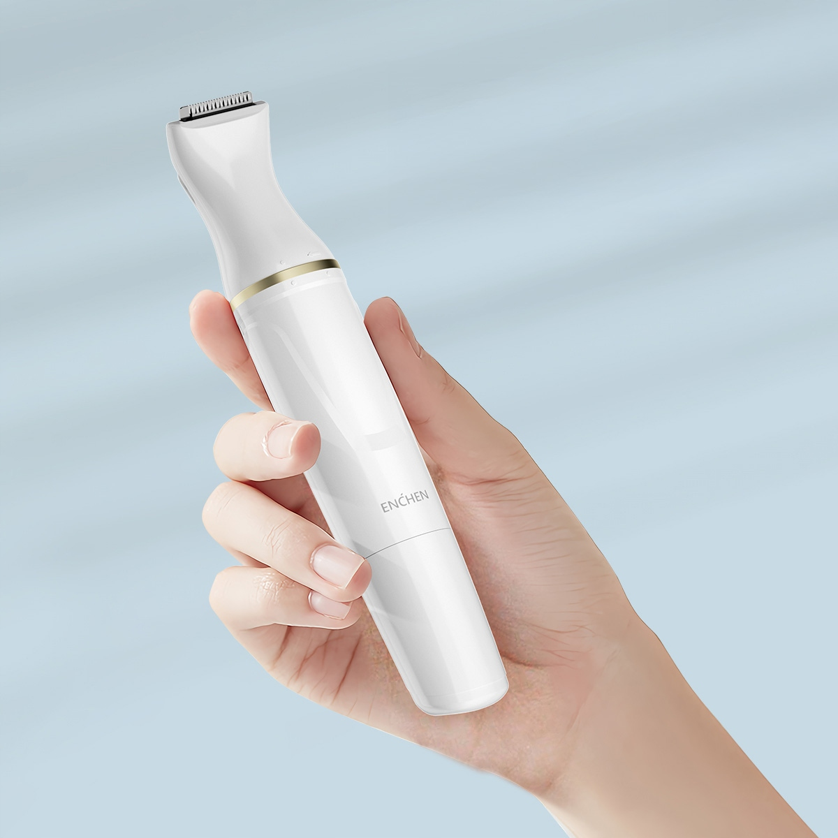 Biała golarka elektryczna trzymana w dłoni na jasnoniebieskim tle. Golarka ma złoty pierścień i markę 'ENCHEN'.