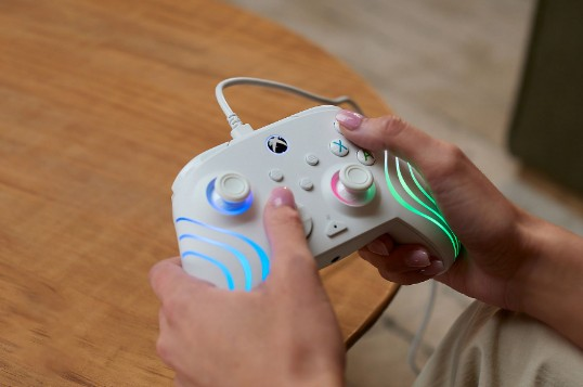 Osoba trzyma biały kontroler Xbox z kolorowymi światłami. Kontroler jest przewodowy i spoczywa na drewnianym stole.