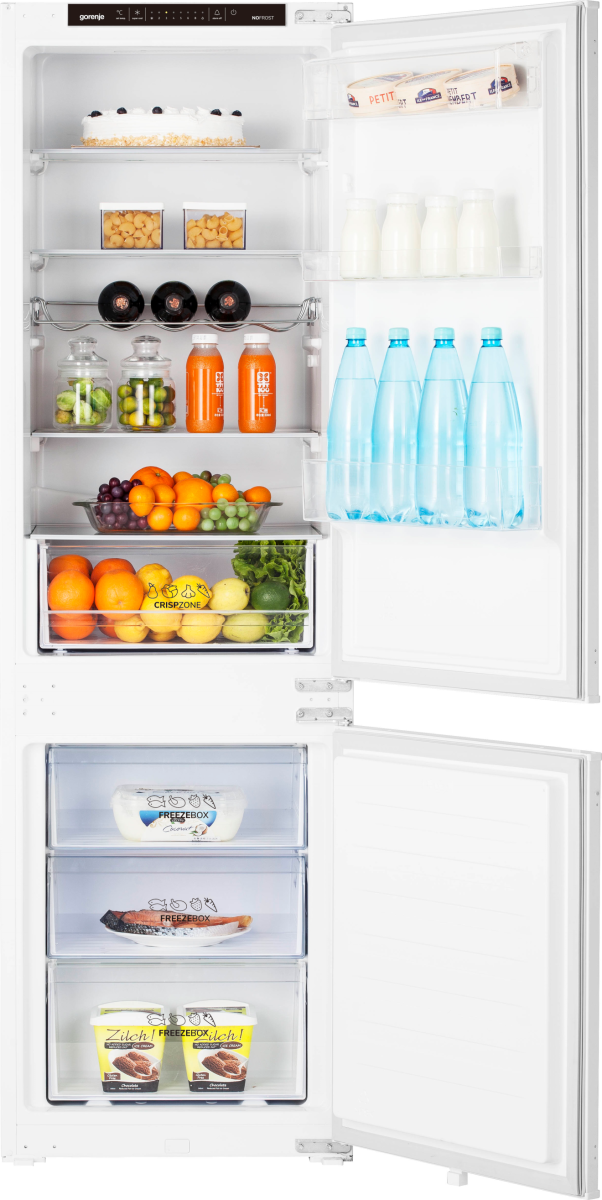 Otwarta lodówka z artykułami spożywczymi: ciasto, butelki, owoce, warzywa i mrożonki. Białe wnętrze z otwartymi drzwiami.