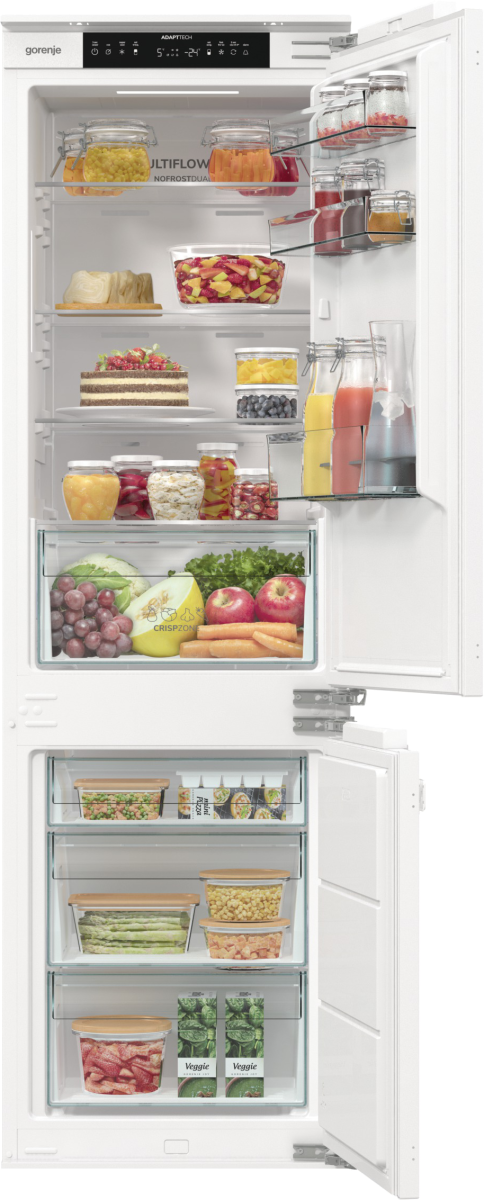 Otwarta lodówka wypełniona jedzeniem: owoce, warzywa, słoiki i pojemniki. Ma wyświetlacz cyfrowy i jest biała.