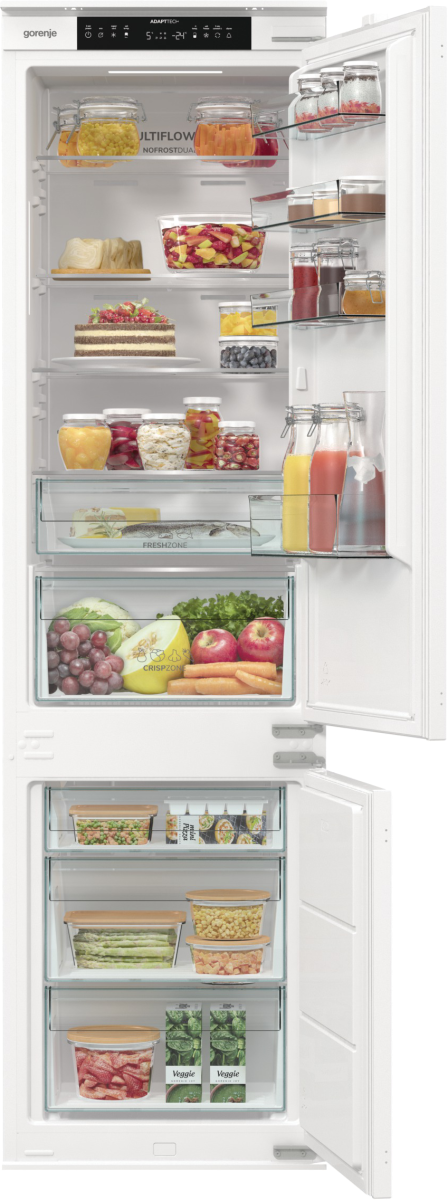 Otwarta biała lodówka wypełniona jedzeniem: słoiki, owoce, warzywa, ciasto i soki. Widoczne półki i szuflady.