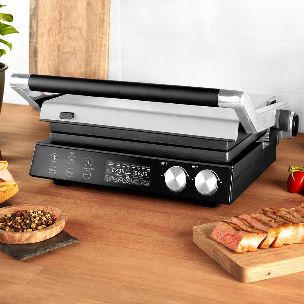 Ein elektrischer Grill mit schwarzem Sockel und silberner Oberseite steht auf einer Holzoberfläche. Steak und Gewürze sind in der Nähe.