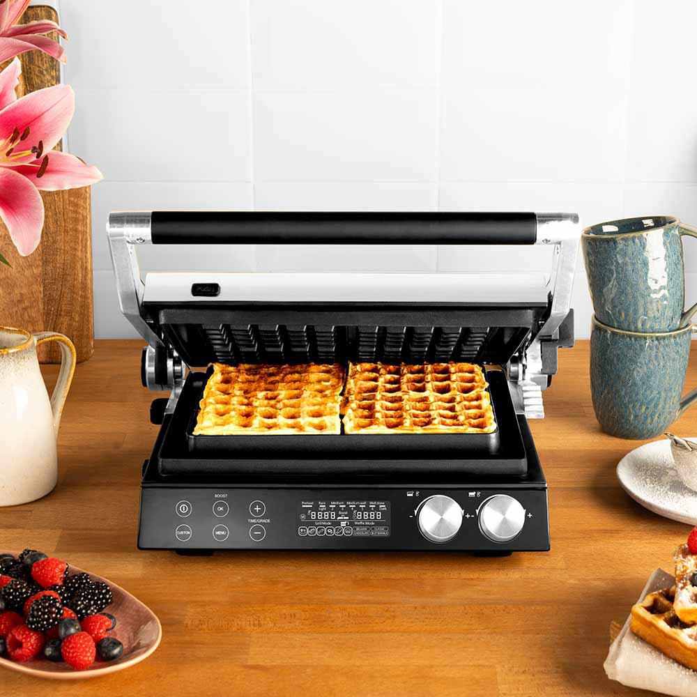 Ein Waffeleisen mit zwei goldenen Waffeln. Beeren, zwei gestapelte Tassen und eine rosa Blume stehen auf Holz.