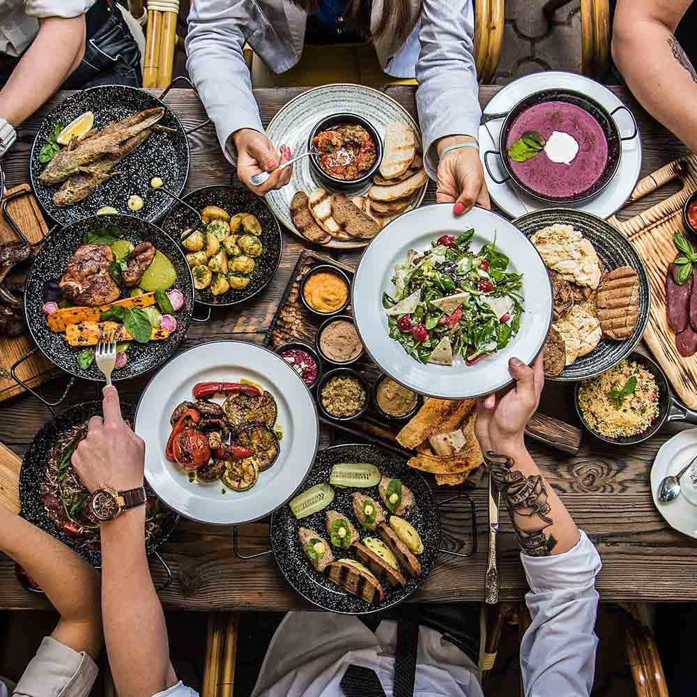 Draufsicht auf einen Tisch mit verschiedenen Gerichten: gegrilltes Fleisch, Gemüse, Salate und Suppen. Mehrere Hände greifen nach dem Essen.