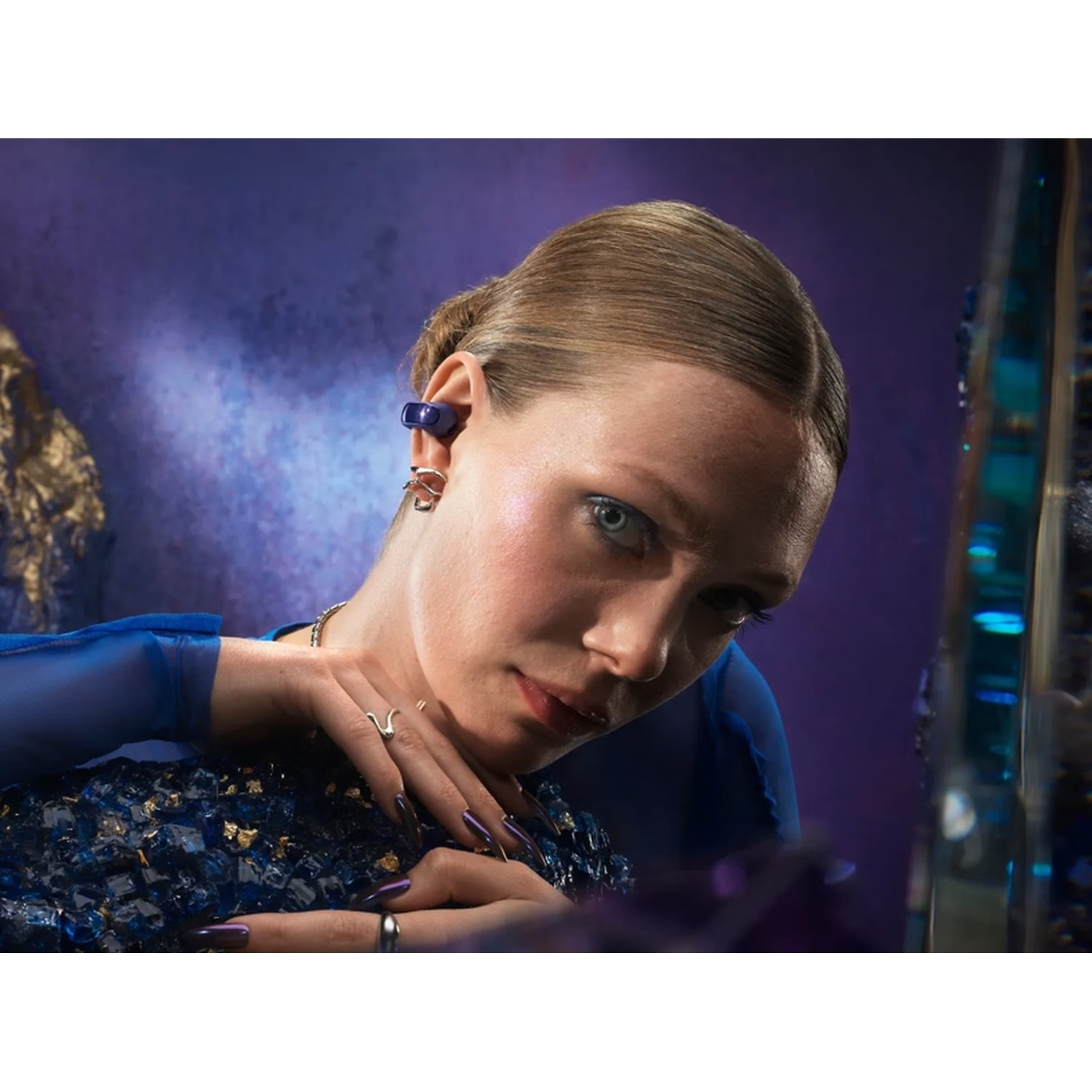 Une femme avec un écouteur dans l'oreille, portant une robe bleue, regardant la caméra. Fond violet foncé et bijoux.