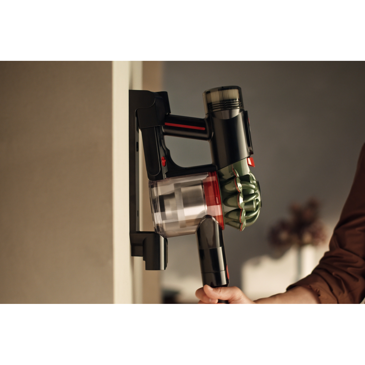 A black and red Dyson vacuum cleaner is mounted on a wall. A hand holds the handle. The wall is white. Background is blurred.
